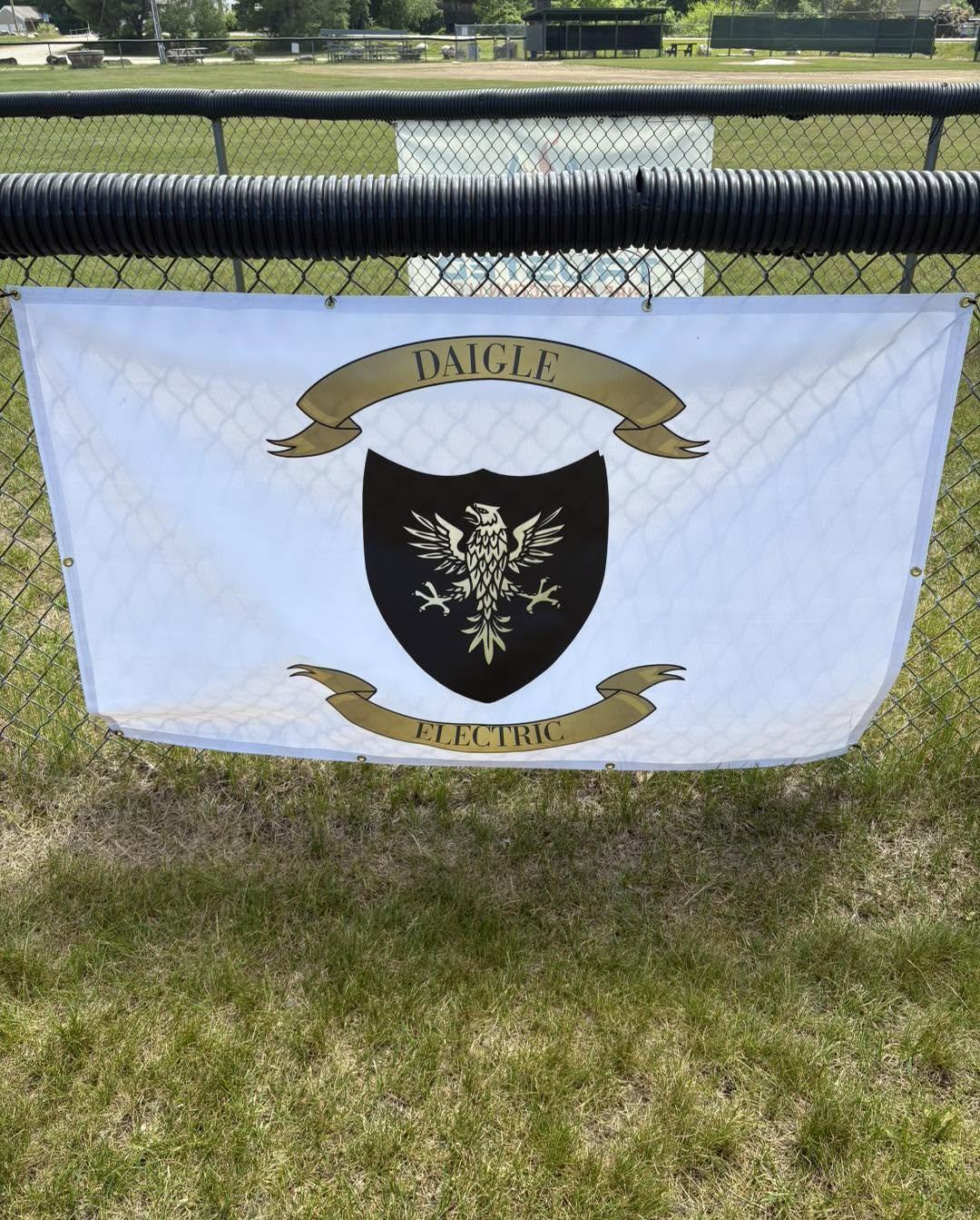 The Daigle Electric banner — white with the black eagle shield — hanging on the outfield fence at the Barrington ballfields.