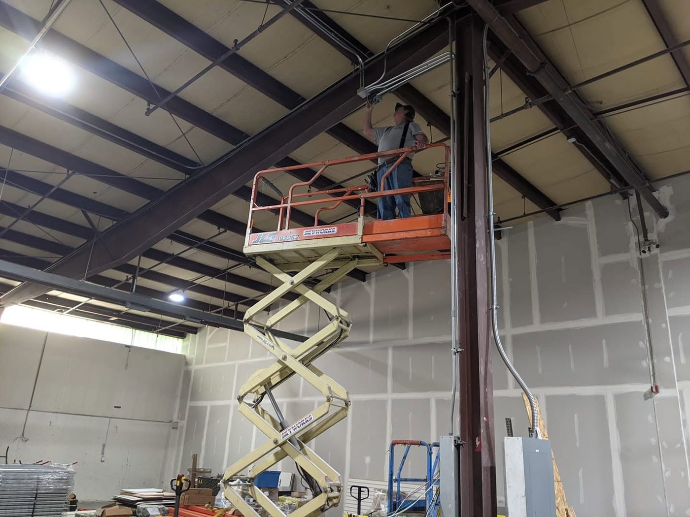 An electrician on a scissor lift running overhead conduit and lighting inside a steel-frame commercial building.