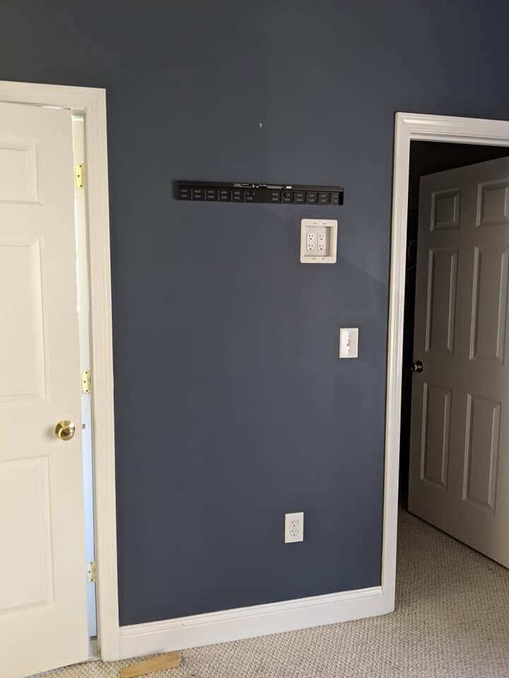 Before: a bedroom wall with a new in-wall outlet and low-voltage plate installed behind a television mounting bracket, with no cables visible on the wall.
