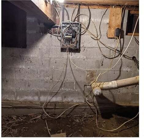 Before: an older undersized panel on a block foundation wall, with branch-circuit cables running loose across the masonry and down onto the basement floor.